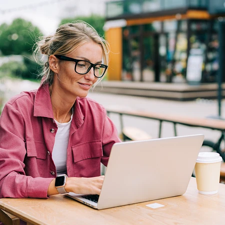 Frau mit Brille arbeitet im Außenbereich eines Cafés an einem Laptop.