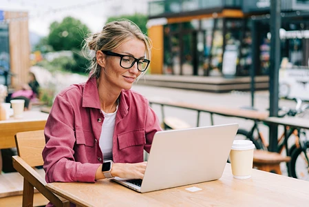 Frau mit Brille arbeitet im Außenbereich eines Cafés an einem Laptop.