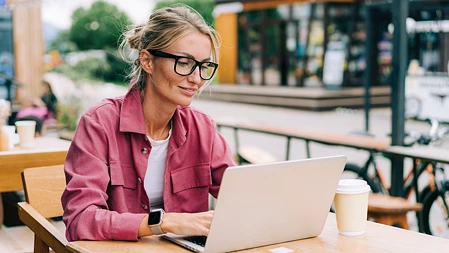 Frau mit Brille arbeitet im Außenbereich eines Cafés an einem Laptop.