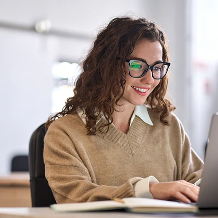 Eine lächelnde Frau mit lockigem Haar und Brille arbeitet an einem Laptop in einem hellen Büro.