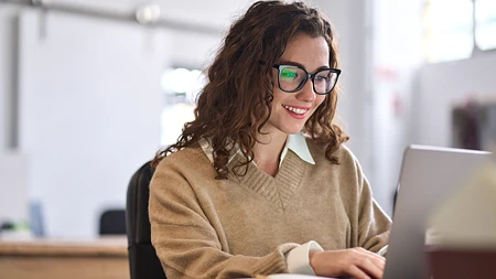 Eine lächelnde Frau mit lockigem Haar und Brille arbeitet an einem Laptop in einem hellen Büro.