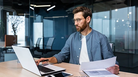 Mann mit Brille und Bart tippt auf einem Laptop in einem Büro