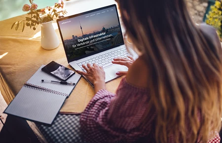 Frau von hinten am Laptop, der eine Website mit dem Text 'Digitale Infrastruktur' zeigt, daneben Notizbuch und Smartphone auf einem Tisch.