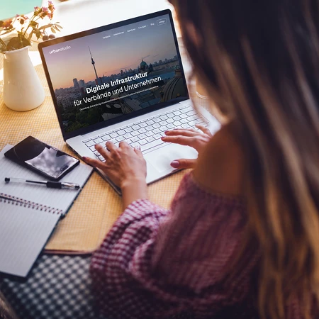 Frau von hinten am Laptop, der eine Website mit dem Text 'Digitale Infrastruktur' zeigt, daneben Notizbuch und Smartphone auf einem Tisch.