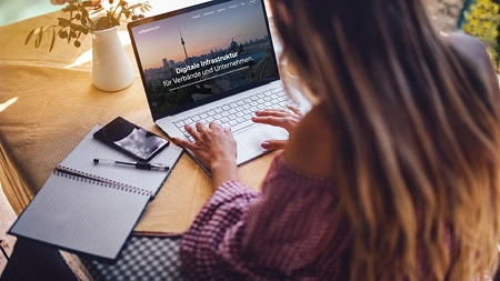 Frau von hinten am Laptop, der eine Website mit dem Text 'Digitale Infrastruktur' zeigt, daneben Notizbuch und Smartphone auf einem Tisch.