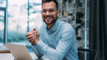 Lächelnder Mann mit Brille und Bart sitzt im Büro vor einem Laptop