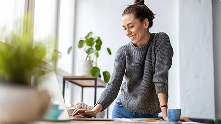 Lachende Frau in grauem Pullover, die an einem Tisch mit Laptop und Tasse arbeitet.