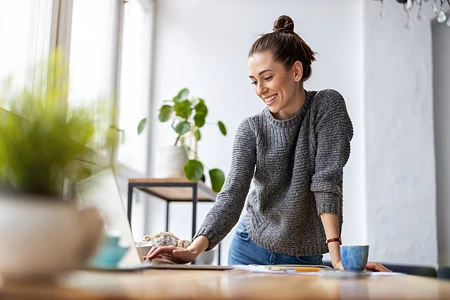 Lachende Frau in grauem Pullover, die an einem Tisch mit Laptop und Tasse arbeitet.
