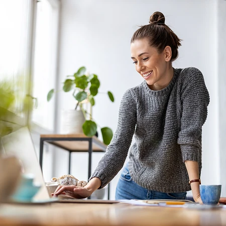 Lachende Frau in grauem Pullover, die an einem Tisch mit Laptop und Tasse arbeitet.