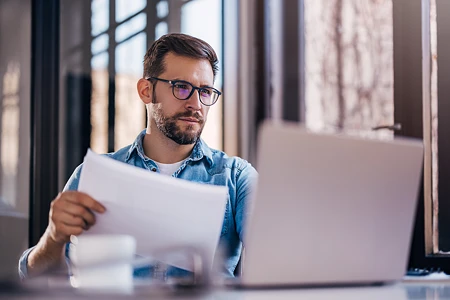 Mann mit Brille und Jeanshemd blickt konzentriert auf einen Laptop und hält ein weißes Blatt Papier. Er prüft, was die Maschine ausgegeben hat.