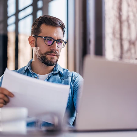 Mann mit Brille und Jeanshemd blickt konzentriert auf einen Laptop und hält ein weißes Blatt Papier. Er prüft, was die Maschine ausgegeben hat.