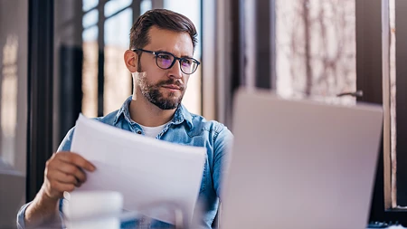 Mann mit Brille und Jeanshemd blickt konzentriert auf einen Laptop und hält ein weißes Blatt Papier. Er prüft, was die Maschine ausgegeben hat.
