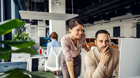 Frau und Mann in Business-Kleidung schauen gemeinsam auf einen Monitor in einem modernen Büro.