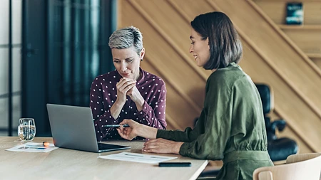 Zwei Frauen in einem Büro, eine zeigt mit einem Stift auf einen Laptop-Bildschirm. Die andere ist offensichtlich skeptisch.
