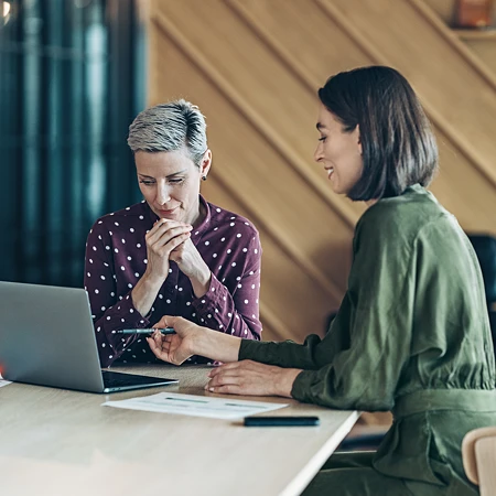 Zwei Frauen in einem Büro, eine zeigt mit einem Stift auf einen Laptop-Bildschirm. Die andere ist offensichtlich skeptisch.