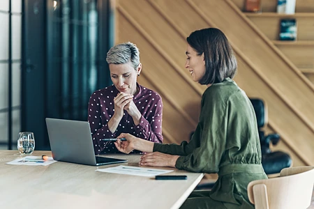 Zwei Frauen in einem Büro, eine zeigt mit einem Stift auf einen Laptop-Bildschirm. Die andere ist offensichtlich skeptisch.