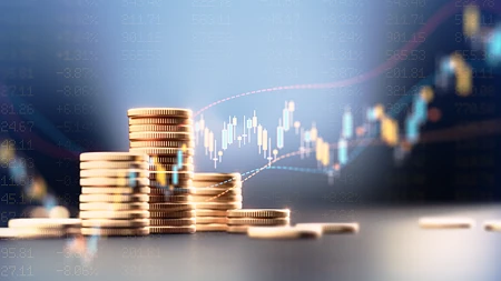 Stapel von Goldmünzen vor einem Hintergrund mit blauem Börsenchart und Finanzdaten.