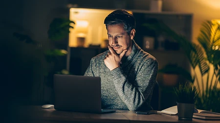 Ein Mann mit nachdenklichem Gesichtsausdruck arbeitet nachts an einem Laptop in einem gemütlich beleuchteten Zimmer.