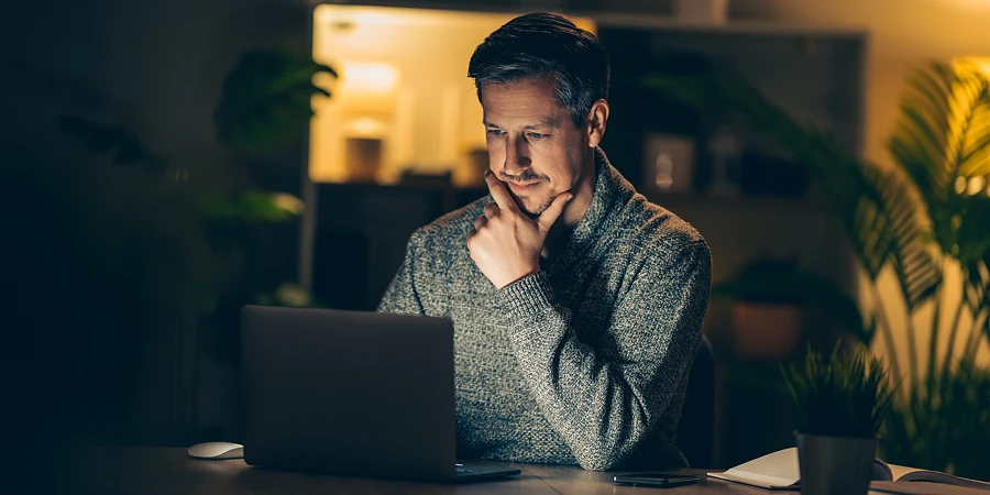 Ein Mann mit nachdenklichem Gesichtsausdruck arbeitet nachts an einem Laptop in einem gemütlich beleuchteten Zimmer.