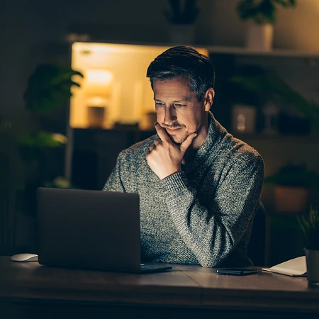 Ein Mann mit nachdenklichem Gesichtsausdruck arbeitet nachts an einem Laptop in einem gemütlich beleuchteten Zimmer.