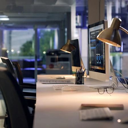 Leeres Großraumbüro bei Nacht, auf einem Schreibtisch leuchtet ein Monitor mit einem Daten-Dashboard, die Stühle davor sind unbesetzt.