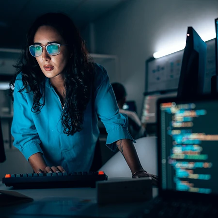 Eine Frau mit Brille arbeitet konzentriert an einem Schreibtisch mit mehreren Monitoren, die Programmiercode anzeigen.