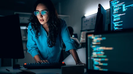 Eine Frau mit Brille arbeitet konzentriert an einem Schreibtisch mit mehreren Monitoren, die Programmiercode anzeigen.