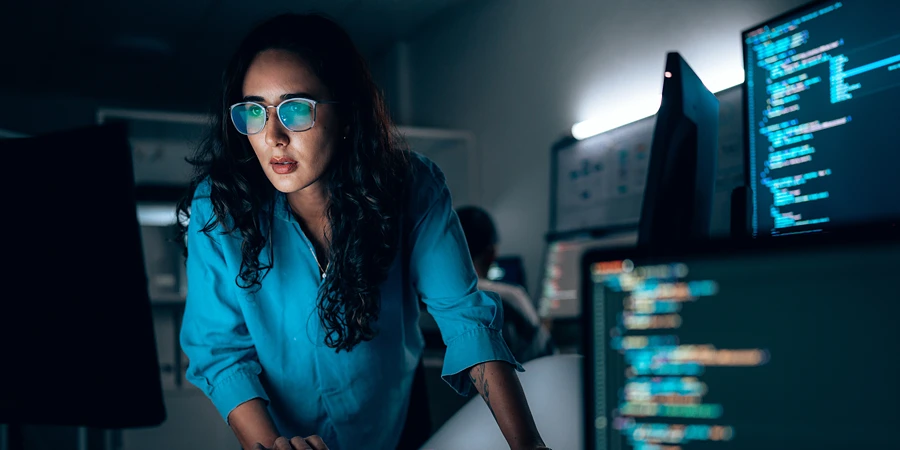 Eine Frau mit Brille arbeitet konzentriert an einem Schreibtisch mit mehreren Monitoren, die Programmiercode anzeigen.